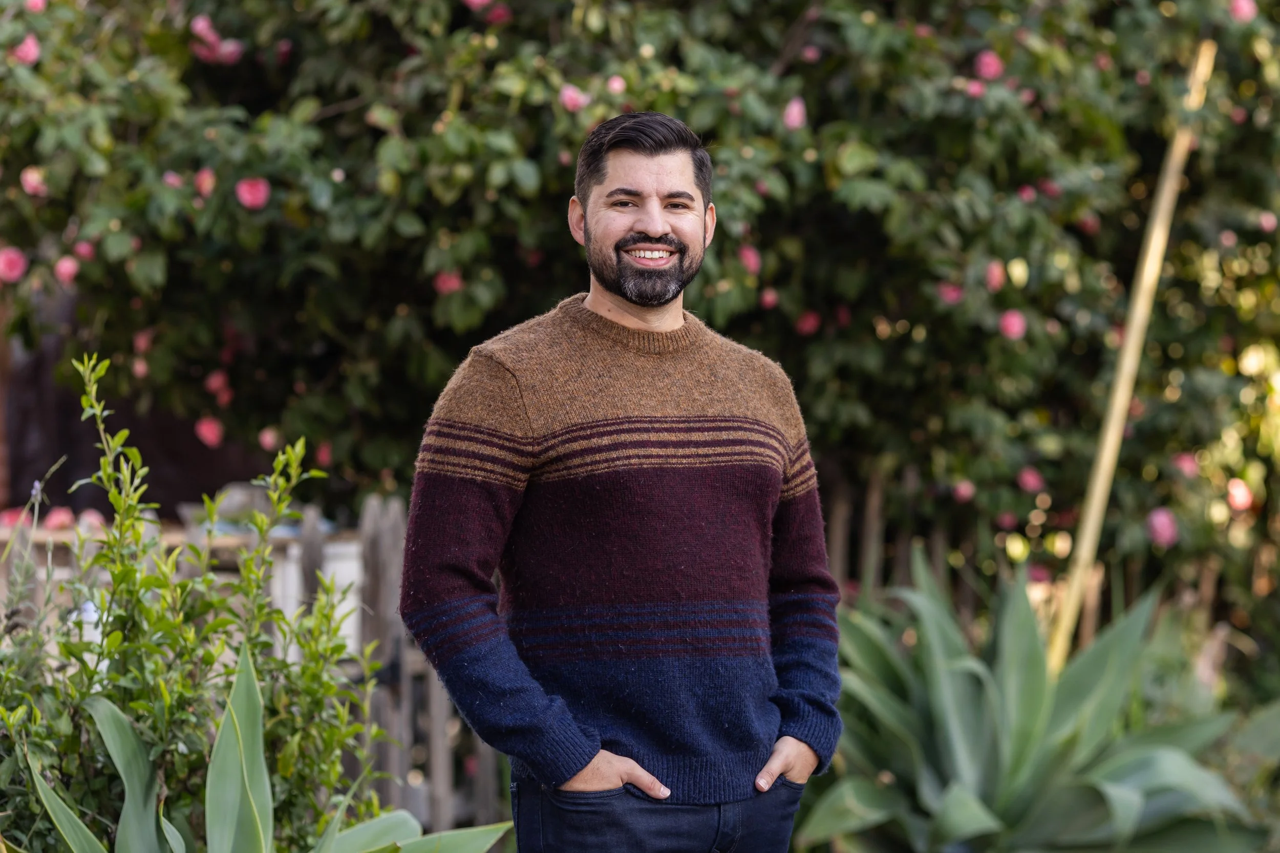 Smiling man with dark hair and beard standing outdoors in front of flowering bushes, wearing a multicolored sweater and blue jeans.