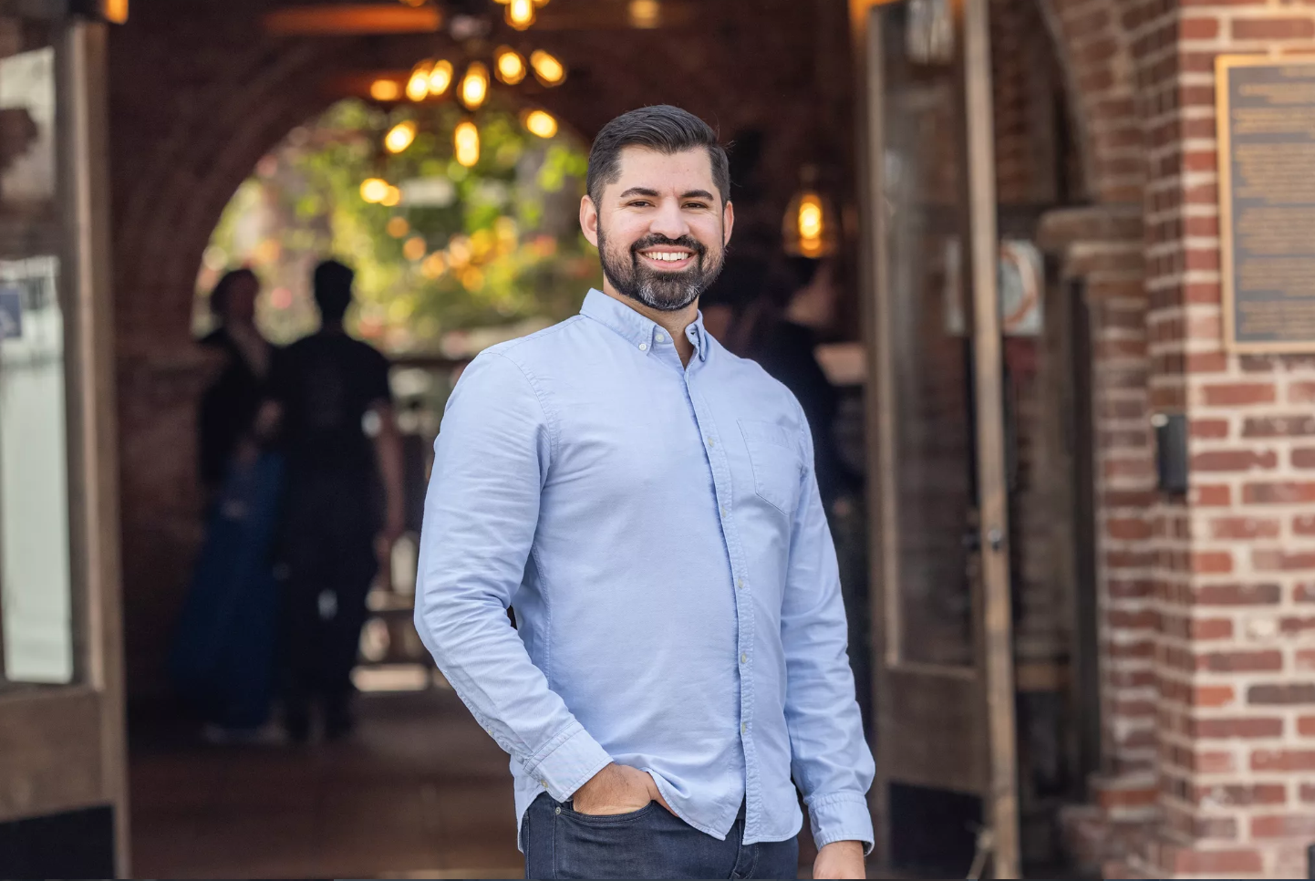 A smiling man with a beard wearing a light blue button-up shirt and dark jeans standing outside in front of a brick building, with people in the background.
